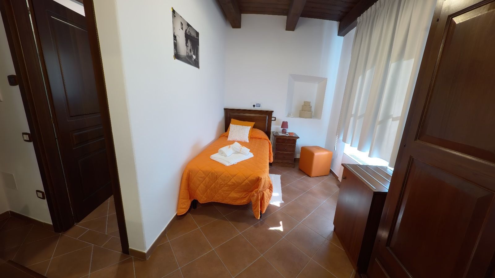 Piccola camera da letto con letto singolo, biancheria arancione, comodino e finestra con tende bianche.