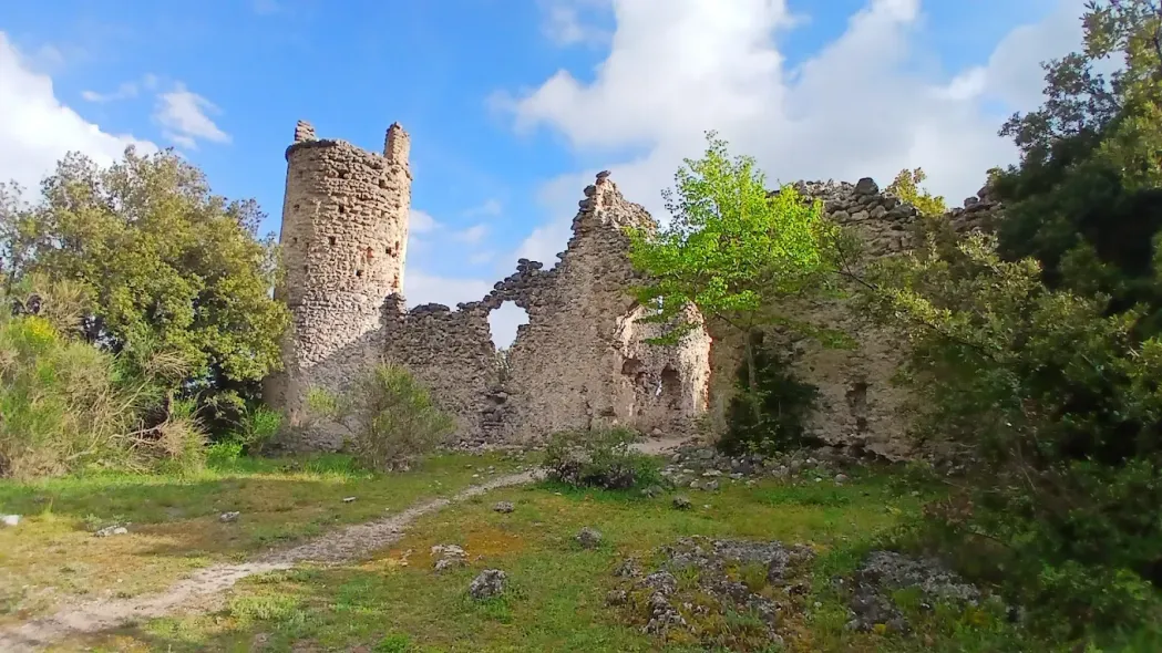 Rovine di un castello in pietra con una torre rotonda, situato su una collina erbosa sotto un cielo azzurro.