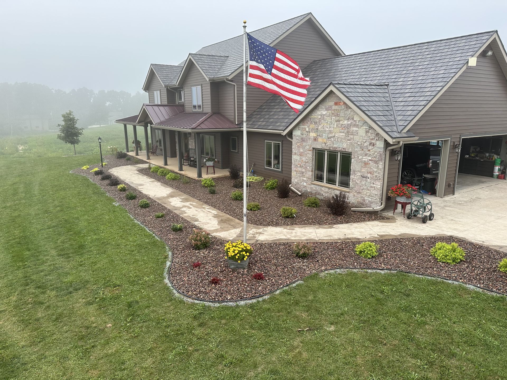 Large stone suburban house with an American flag, front porch, and landscaped lawn in foggy weather