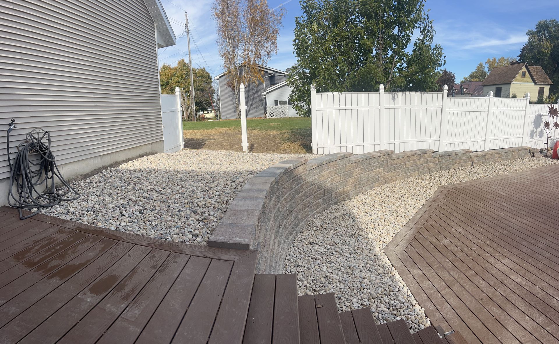 Backyard with gravel path, wooden deck, white fence, and house exterior under a blue sky