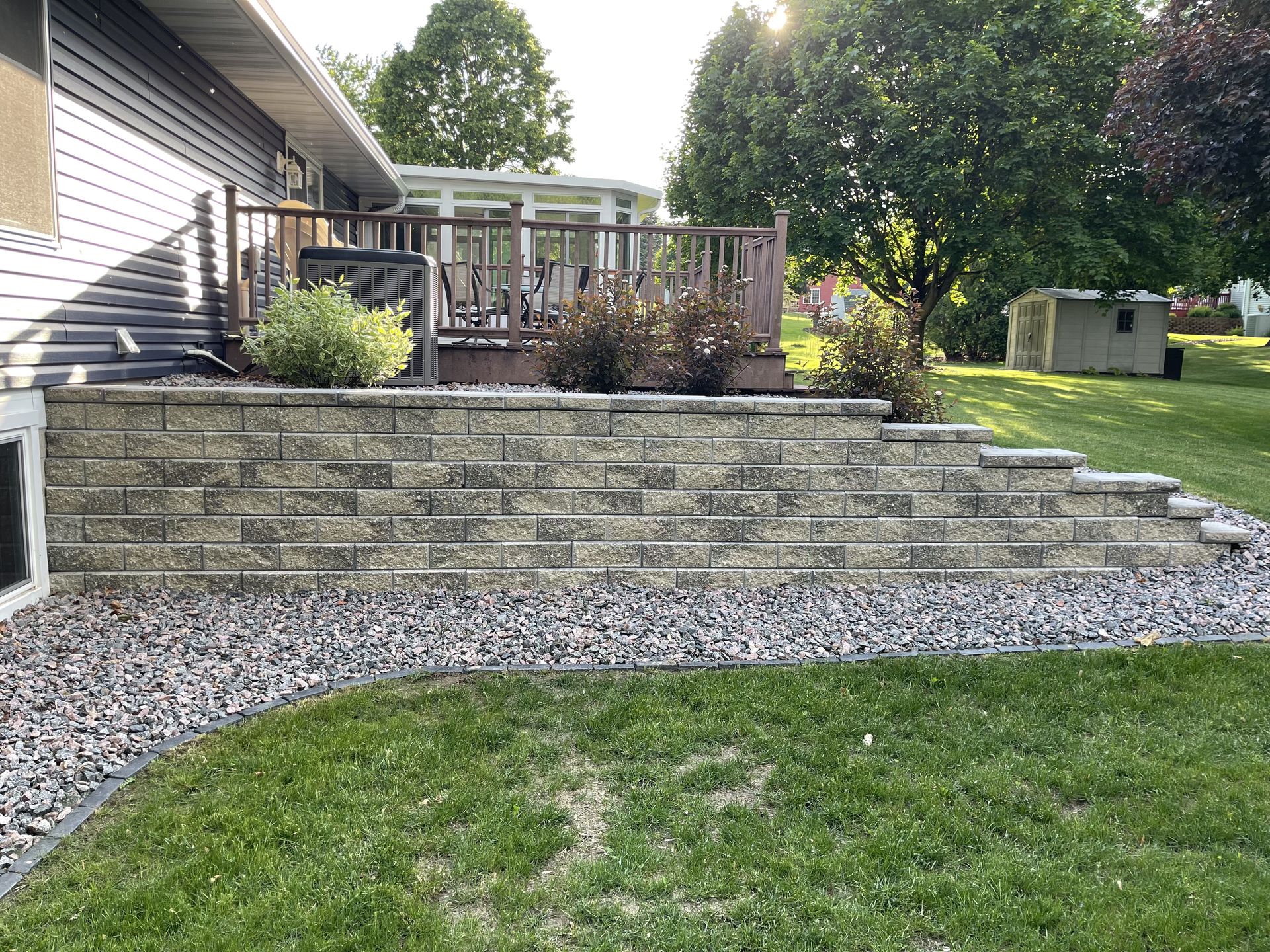 Backyard patio with stone retaining wall, wooden deck, gravel border, and green lawn