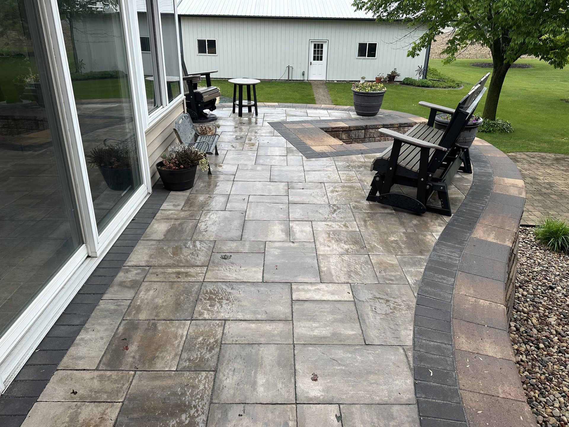 Stone patio with outdoor chairs and a fire pit beside a house, bordered by mulch and a lawn.