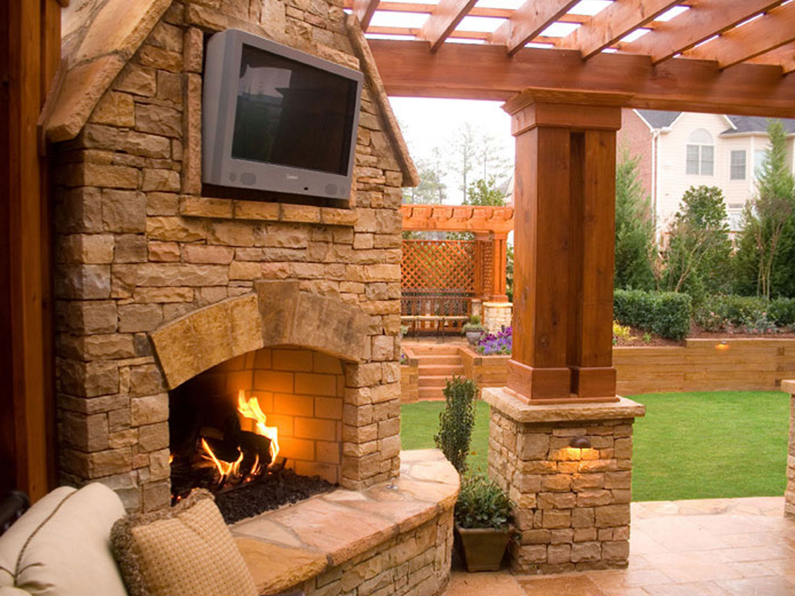 Stone outdoor fireplace under a wooden pergola with a burning fire and TV above it, overlooking a landscaped yard