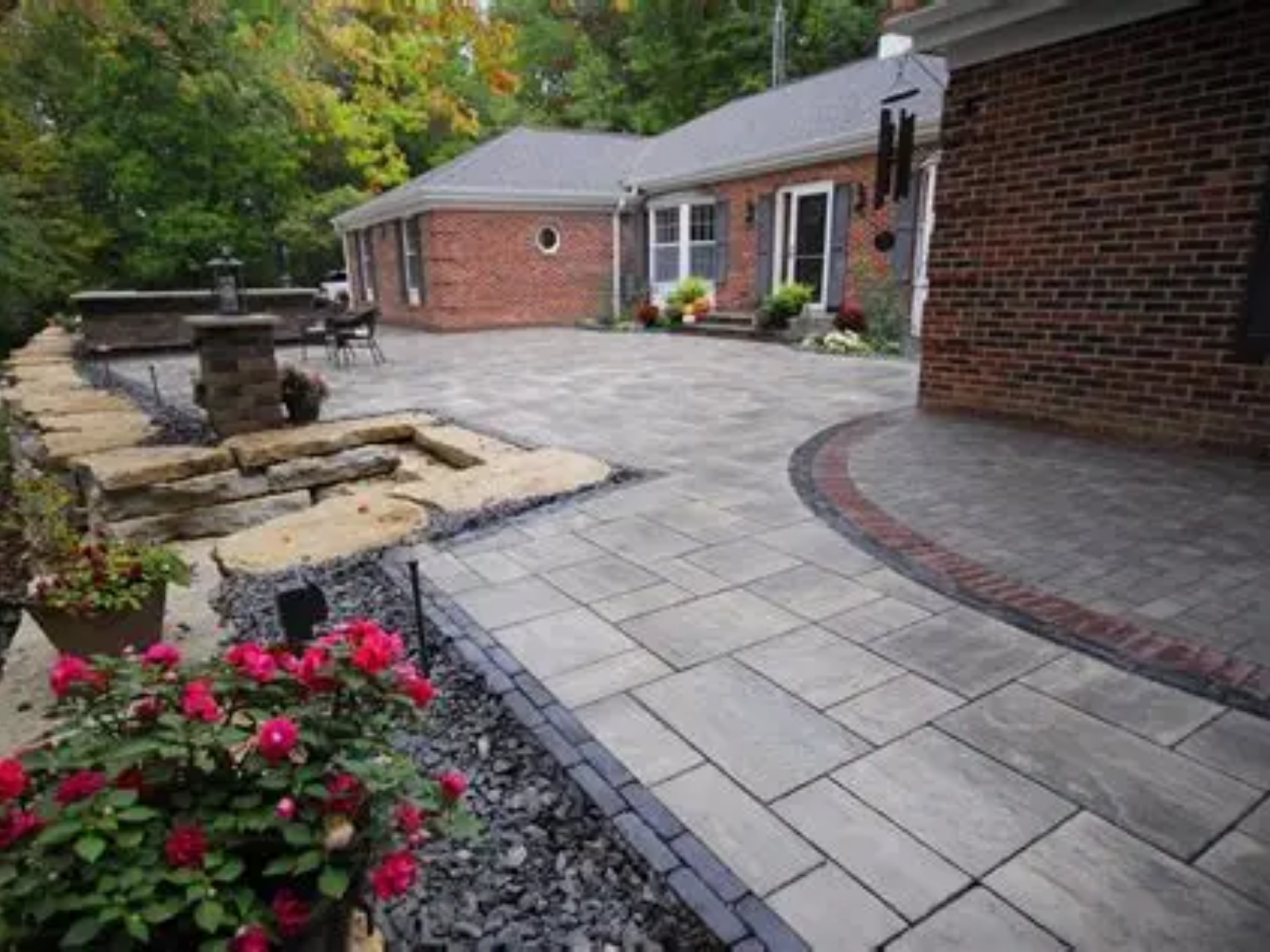 Stone patio and curved walkway beside a brick house, with a small pond and pink flowers.