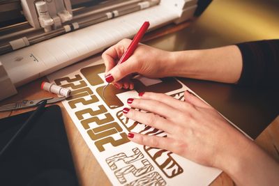 Hands writing on a sheet of gold stickers with a red pen, next to a cutting machine.