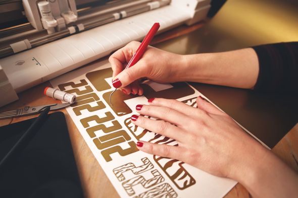Hands writing on a sheet of gold stickers with a red pen, next to a cutting machine.