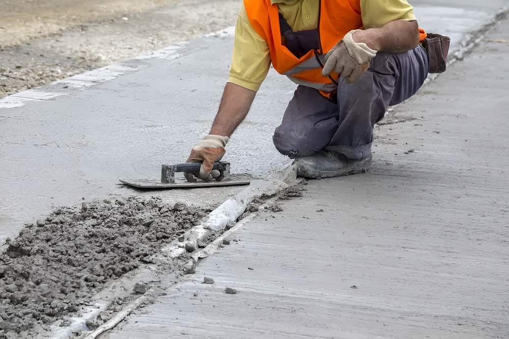 A Man Is Kneeling Down And Using A Trowel To Spread Concrete On A Sidewalk — Ingham Concrete–Icon in Ingham, QLD