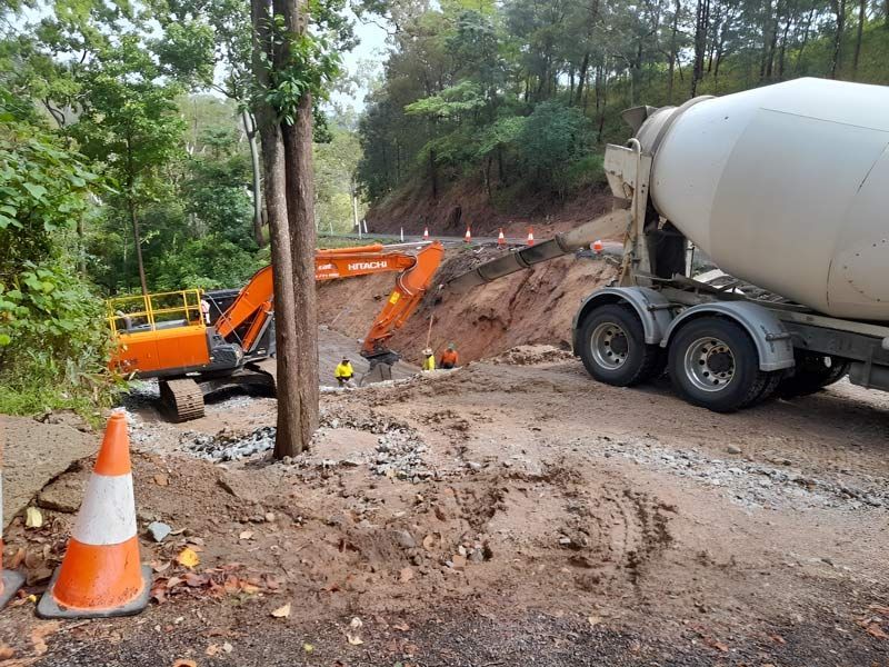 A Concrete Mixer Truck Is Driving Down A Dirt Road Next To An Excavator — Ingham Concrete–Icon in Ingham, QLD