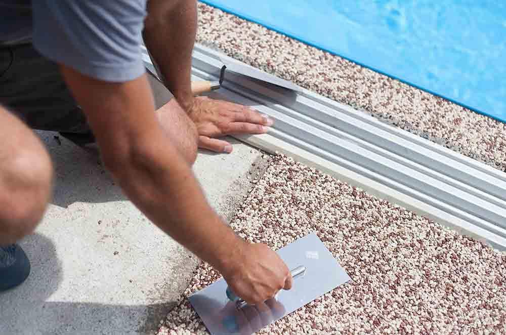 Man Is Kneeling Down And Using A Trowel On A Concrete Floor Next To A Pool — Ingham Concrete–Icon in Ingham, QLD
