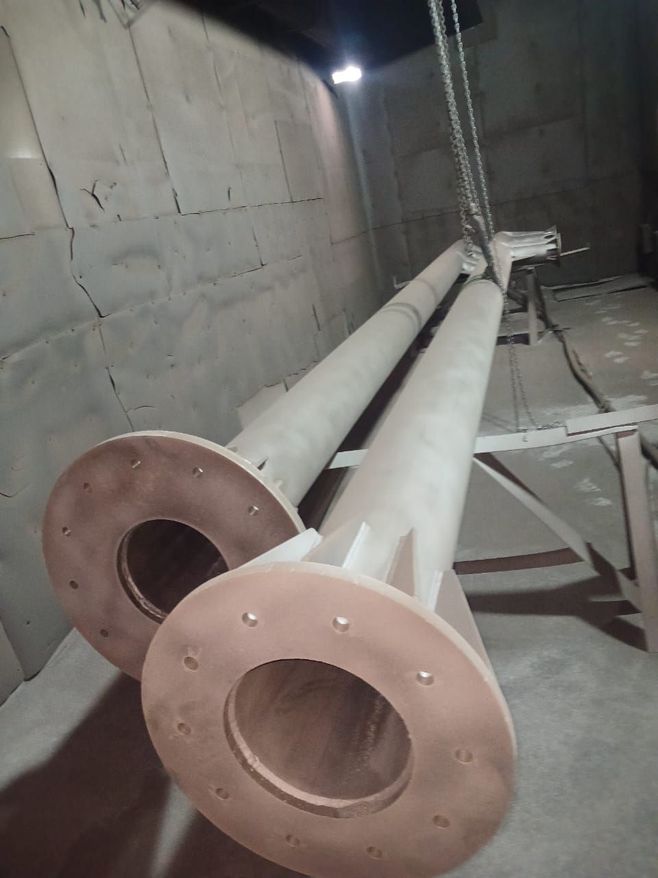 Dos largos postes metálicos de color beige, con bridas circulares, colgados en un espacio industrial poco iluminado.