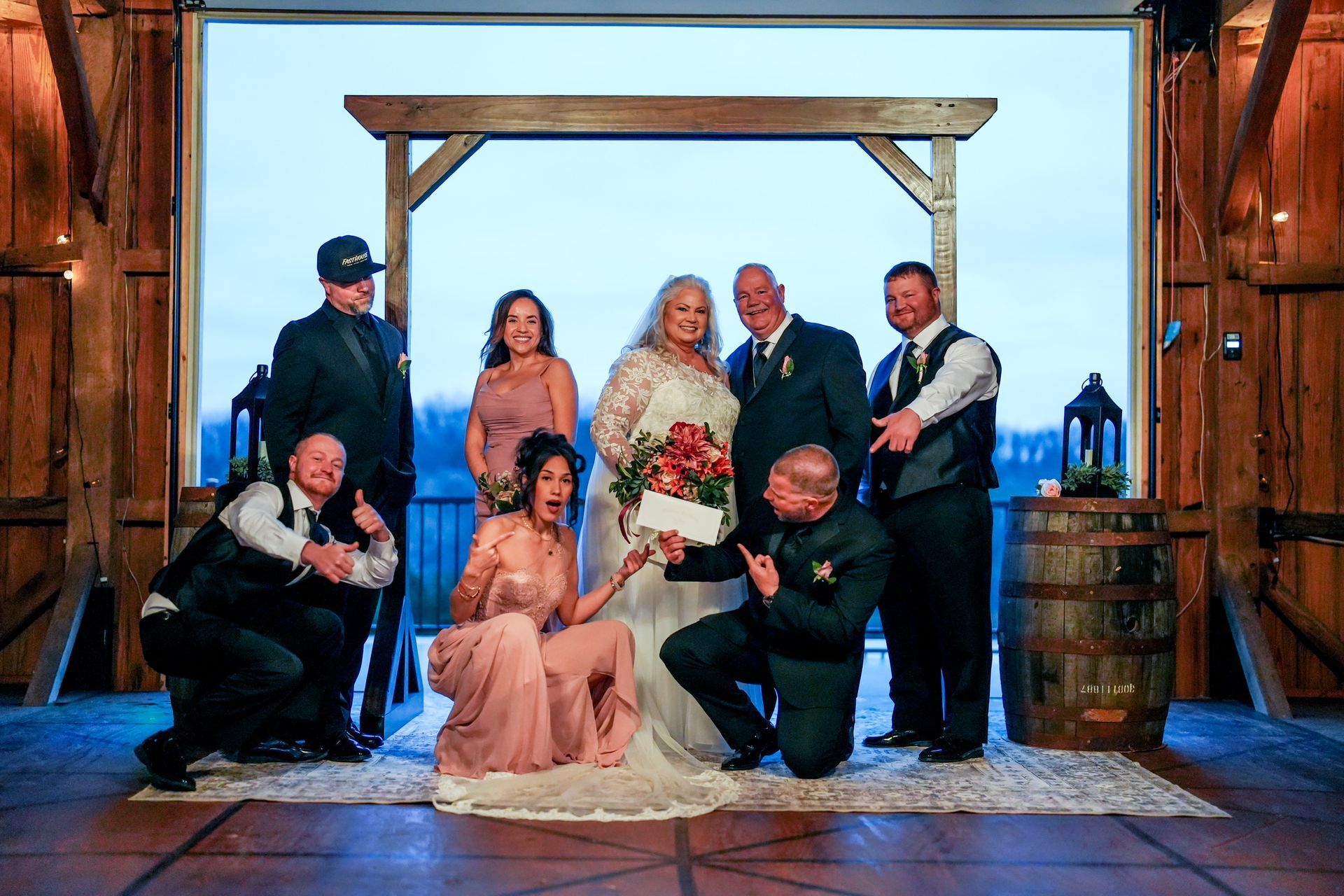 Wedding group inside a barn, posing with the bride holding flowers, some people are kneeling.