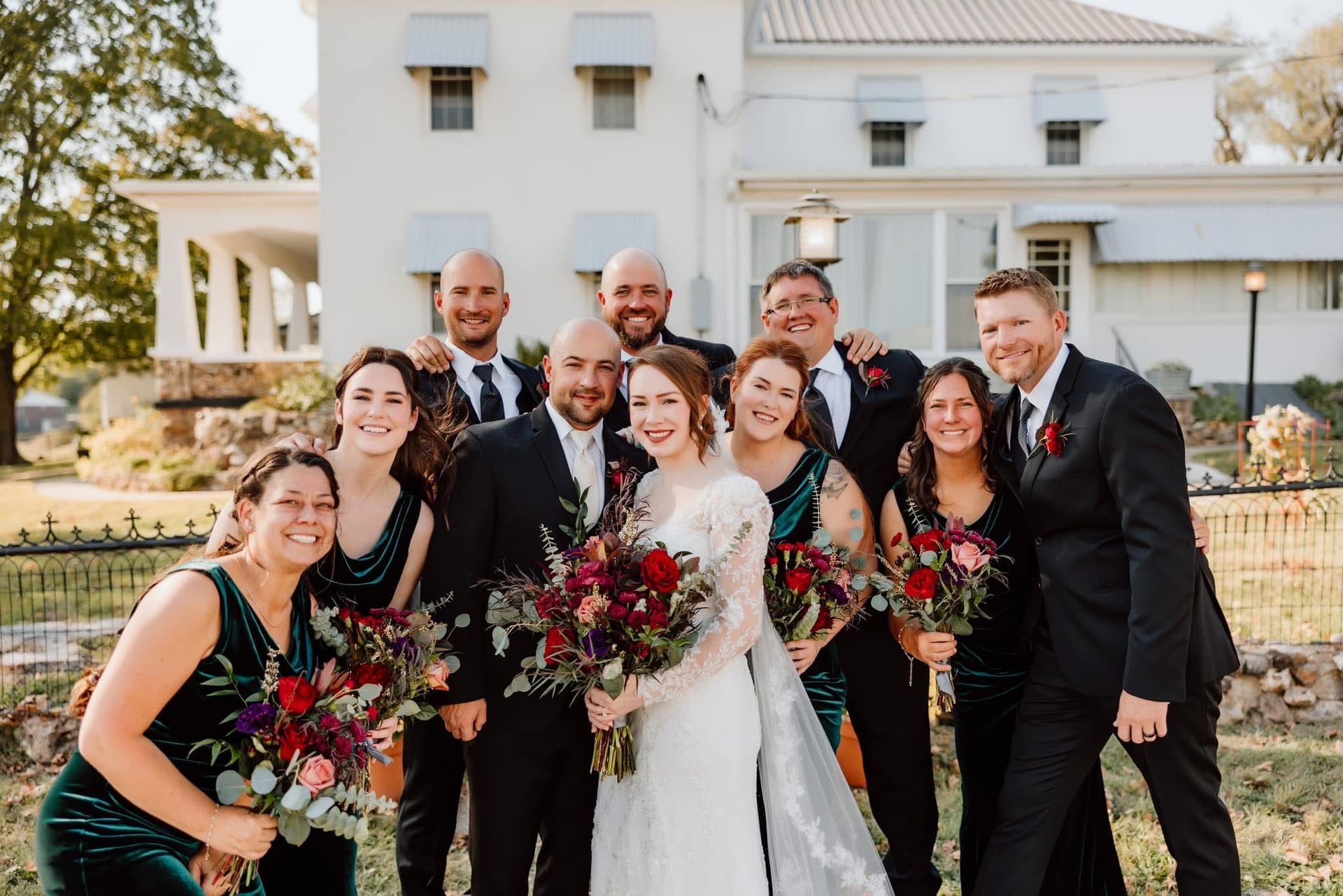 Wedding party posing outside a white building. Bride and groom centered, surrounded by bridesmaids and groomsmen holding bouquets.
