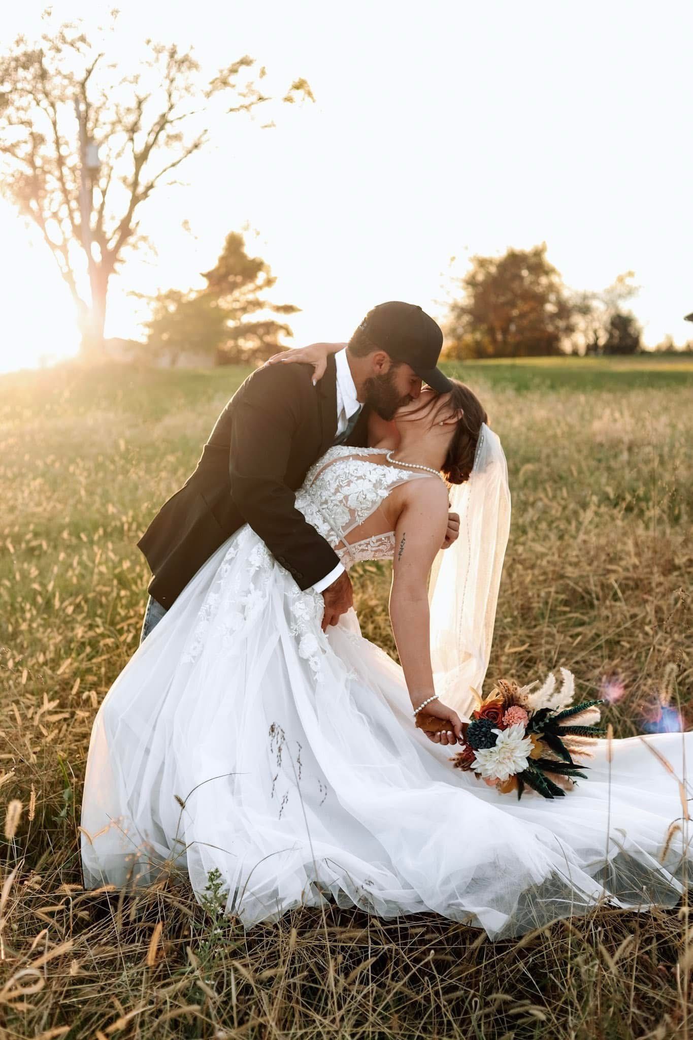 Couple embracing and kissing outdoors on wedding day; woman in white gown, man in black suit, field setting.