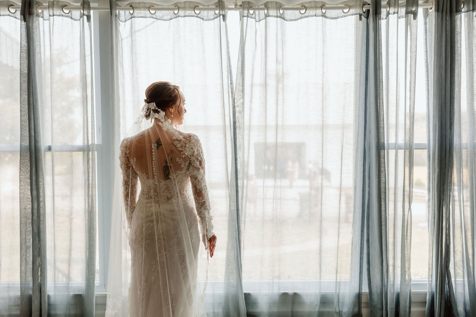 Bride in lace gown looks out a window. She has updo and veil, curtains frame her.