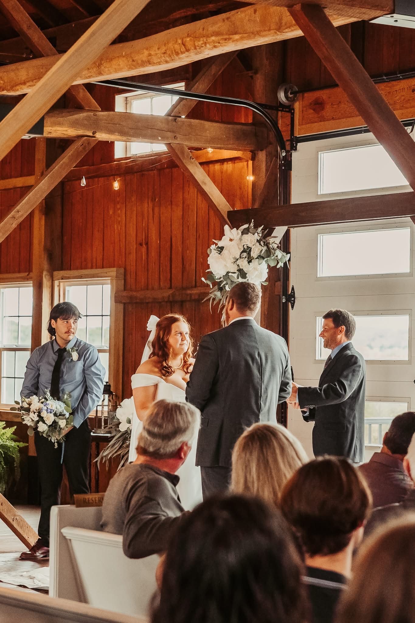 Wedding ceremony inside a barn. Bride and groom face officiant; guests seated. Rustic wood beams and white floral arrangements.