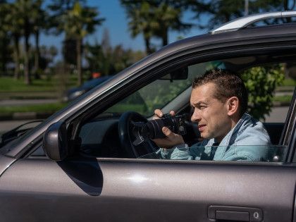 Man in a car taking a photo with a camera. Setting is outdoors, trees in background.