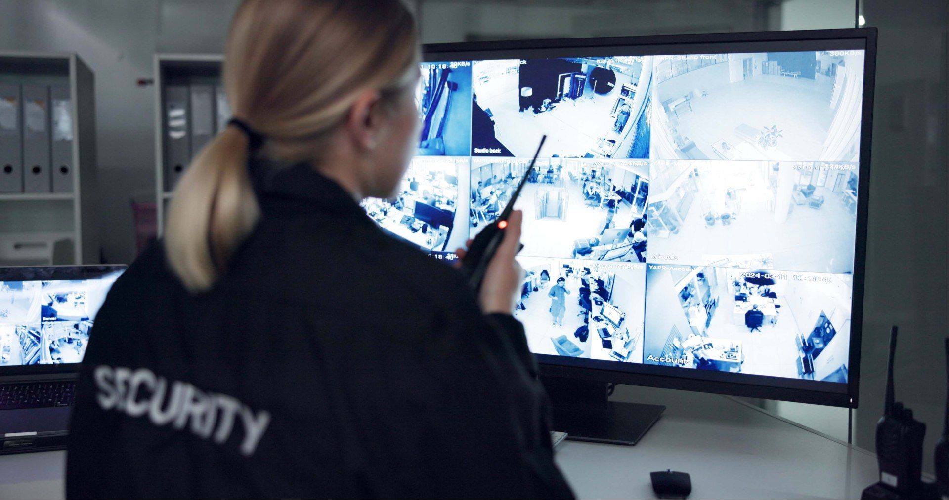 Woman security guard in a control room, looking at multiple surveillance camera views on a monitor while holding a radio.