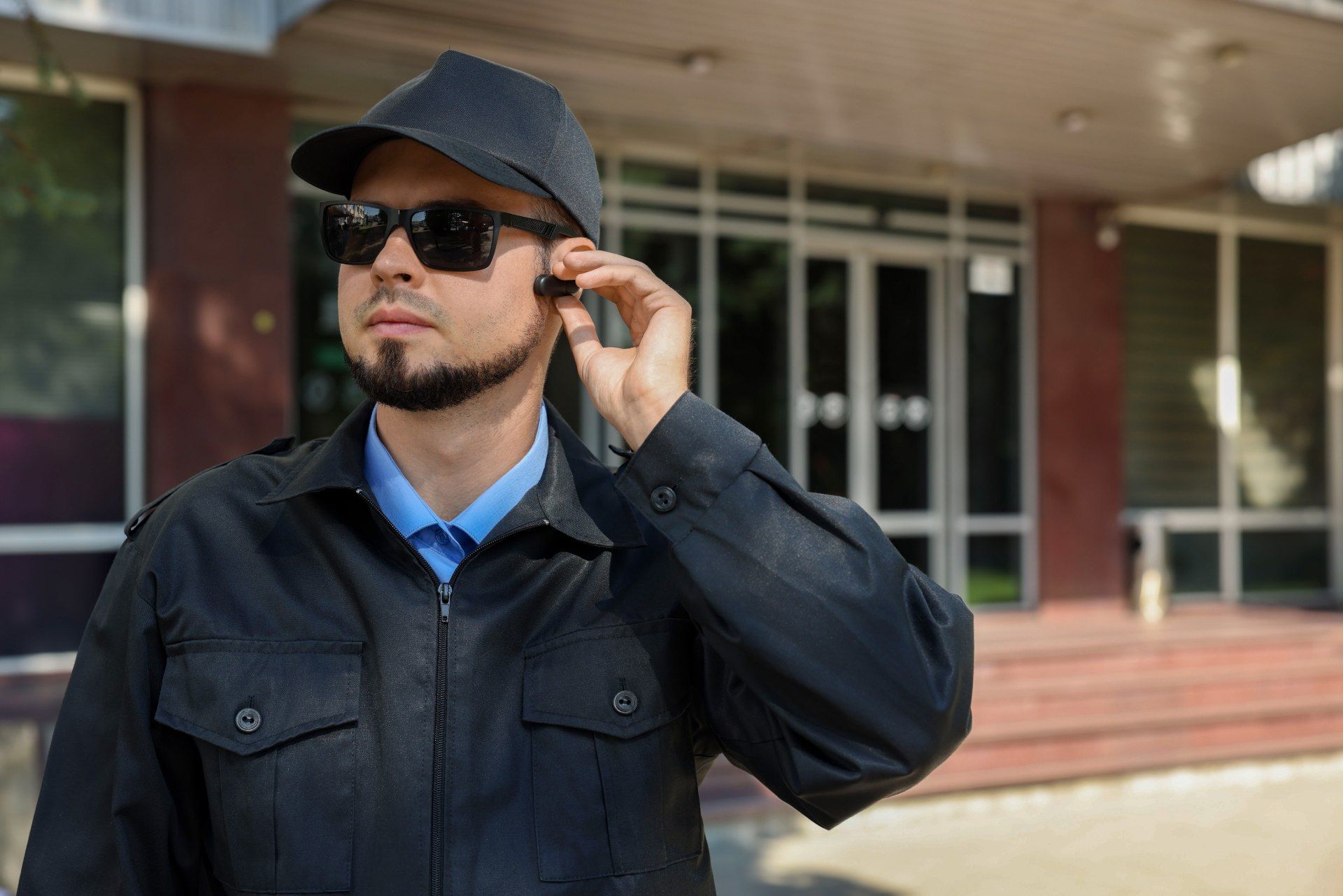 Security guard wearing sunglasses and earpiece, outside building with glass doors.