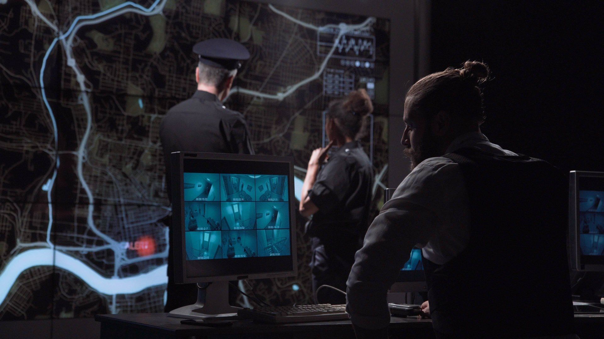 People in a dark control room monitor screens displaying maps and security camera feeds.