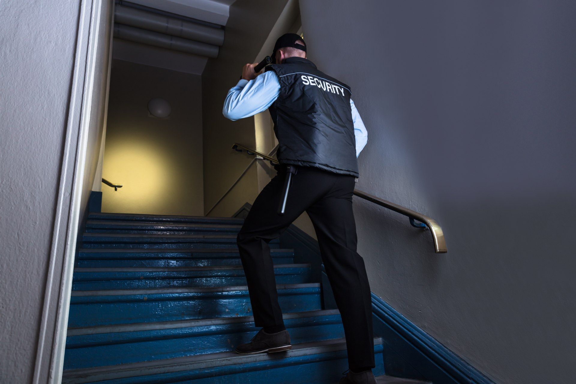Security guard in black uniform climbing blue stairs, speaking into radio, illuminated by a spotlight.