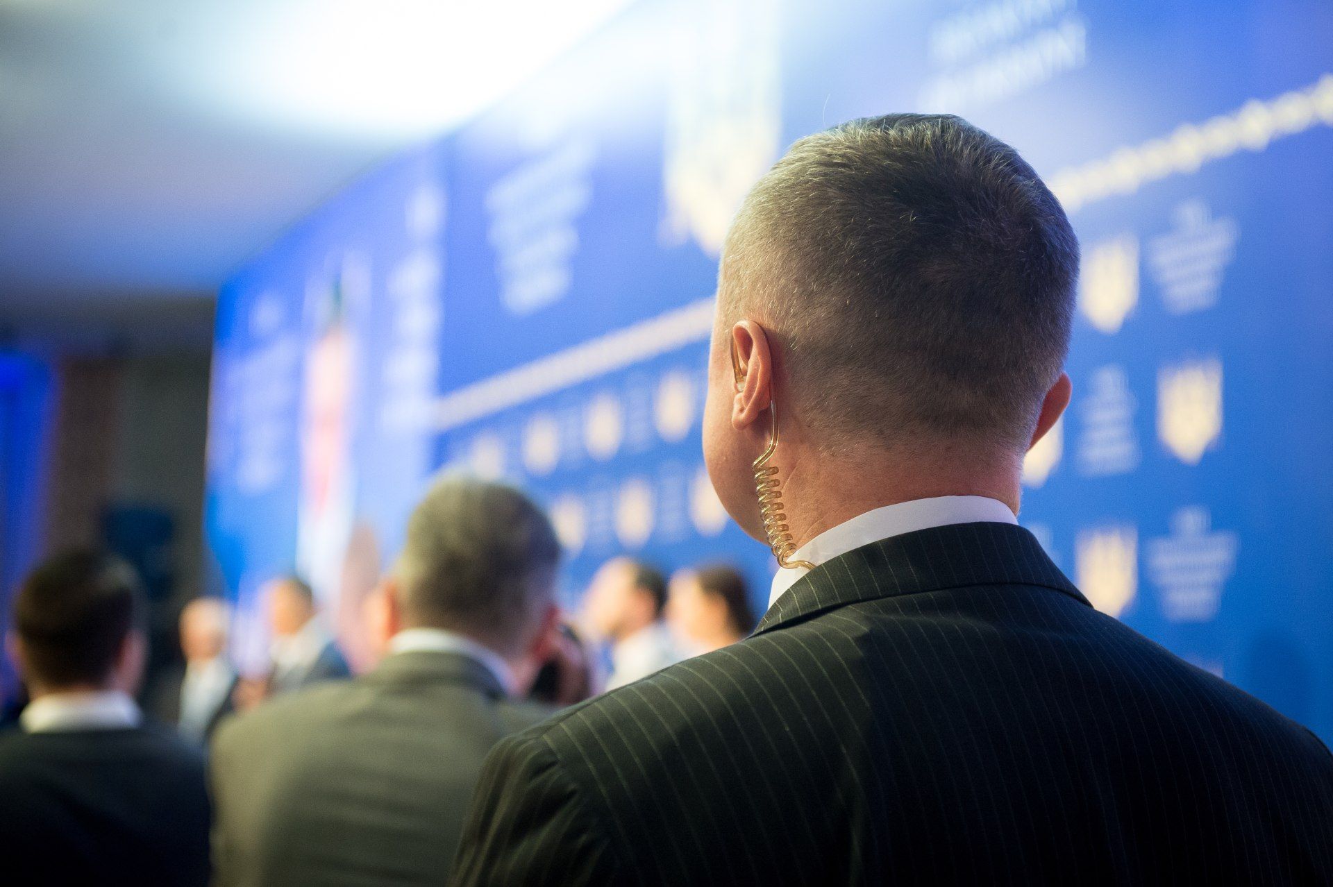 A man in a suit, wearing an earpiece, stands guard at an event with a blue backdrop.