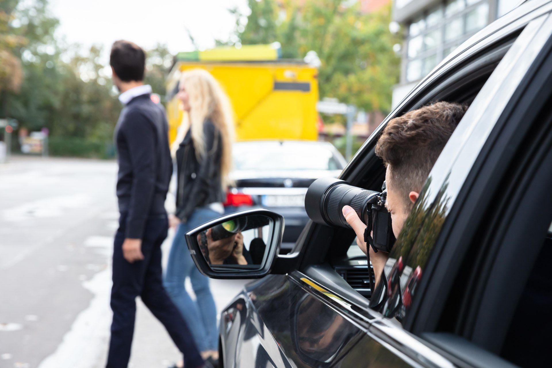 A person in a car taking a photo of a couple walking on a sidewalk with a long lens camera.
