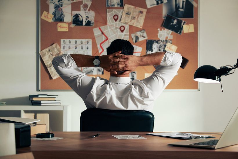 Man in white shirt at desk, hands behind head, gazing at detective's bulletin board in an office.