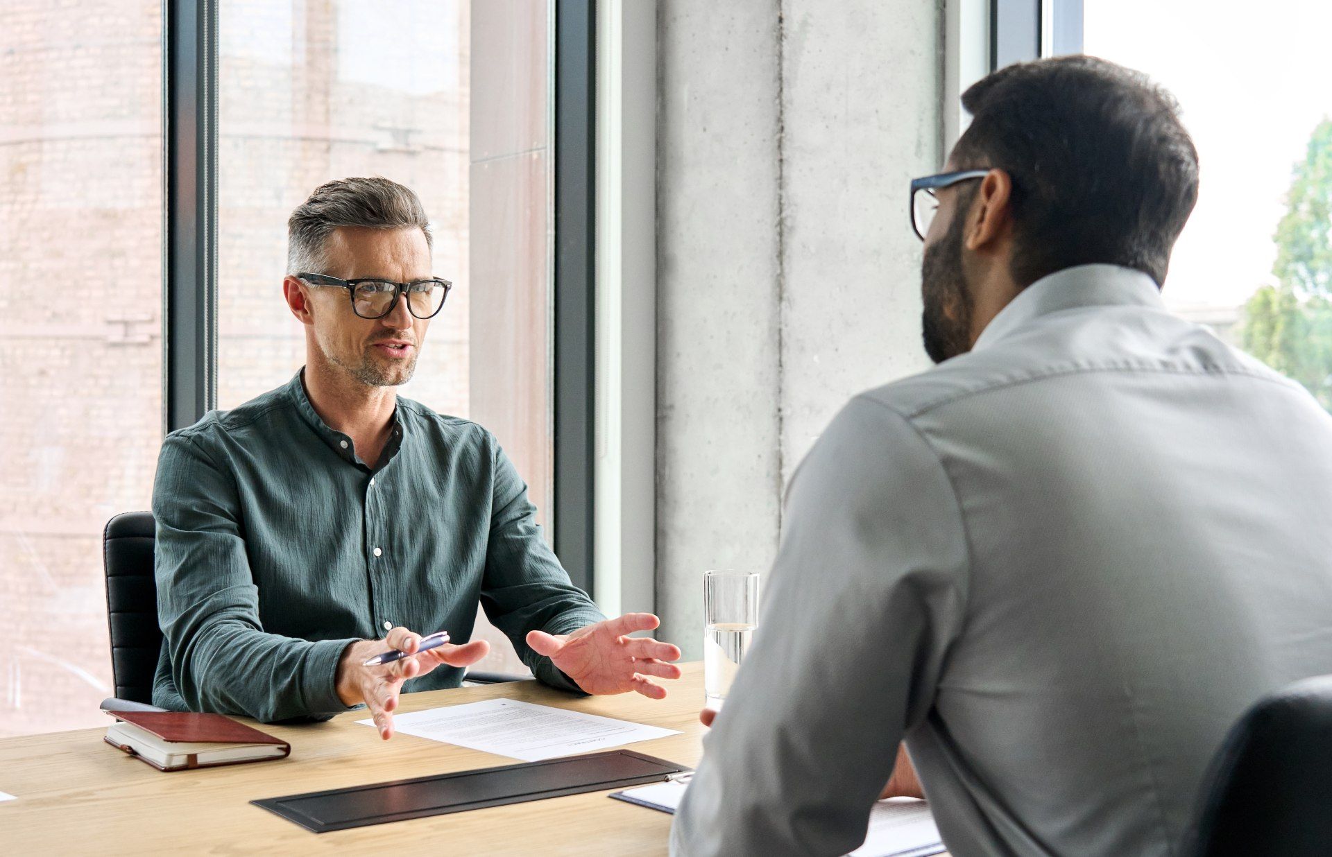 Two men at a desk in an office setting, one gesturing while speaking, the other listening attentively.