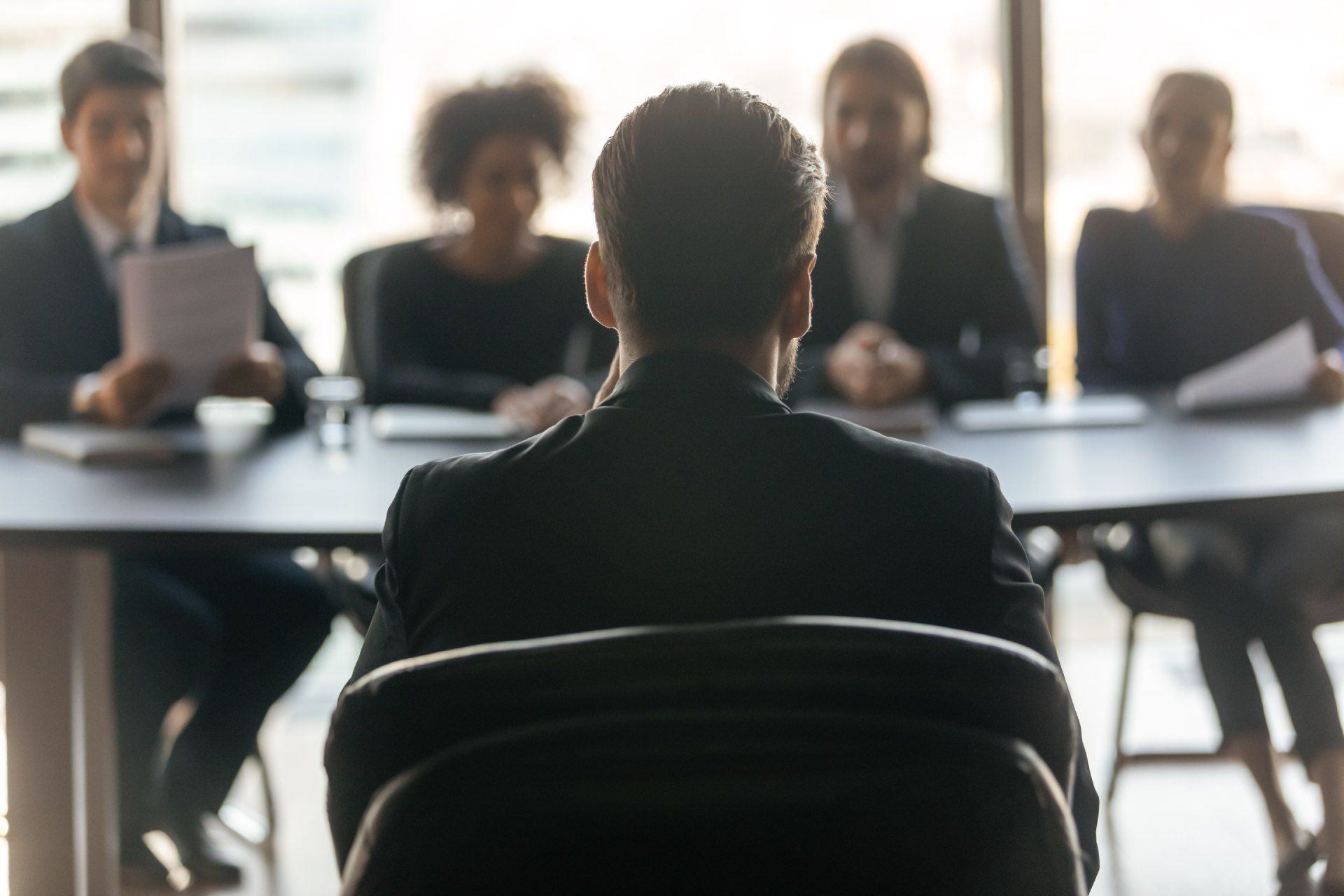 Person facing a panel of interviewers in a bright office setting. Back of interviewee visible.