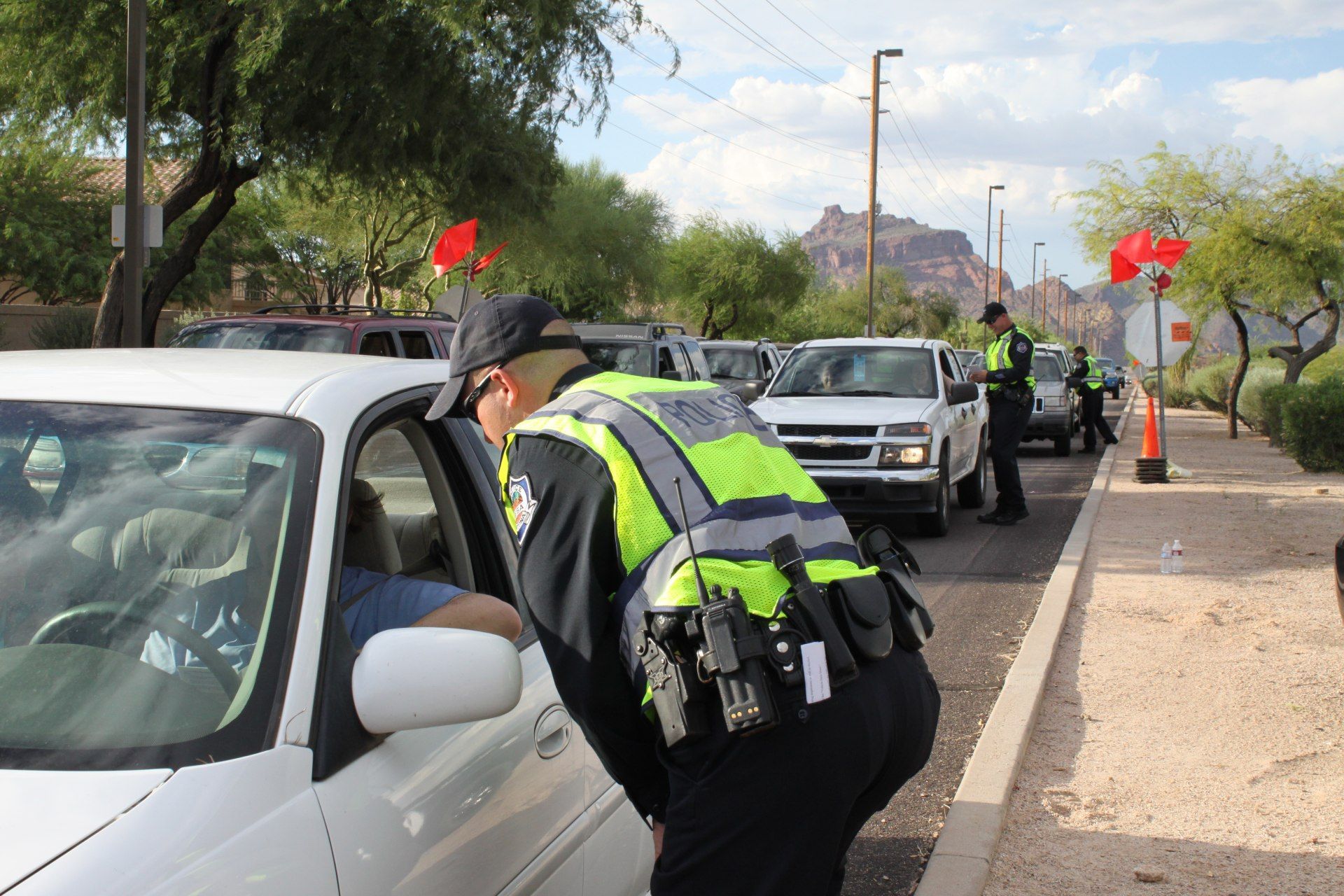 Police officer leaning into car window, speaking to driver at a checkpoint with other vehicles. Outdoor, bright daylight.