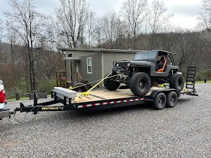 A black Jeep with large off-road tires secured by yellow straps onto a flatbed utility trailer.