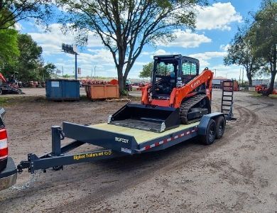 An orange compact track loader sits secured on a flatbed trailer being towed in an outdoor equipment yard.