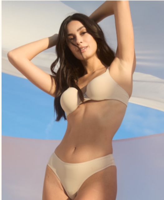 Una donna in lingerie beige posa con le braccia alzate, tenendo in mano un pezzo di stoffa su uno sfondo blu e bianco.