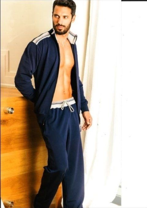 Uomo in tuta blu, con la cerniera abbassata, appoggiato a un mobiletto di legno vicino a una finestra con tenda bianca.