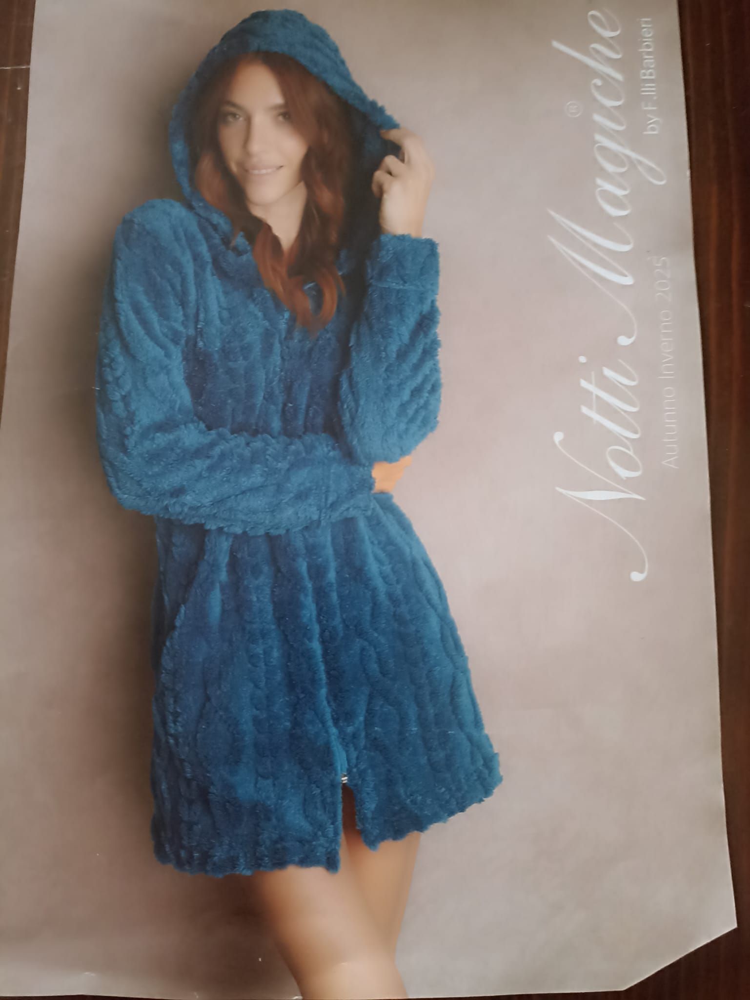 Donna in vestaglia verde acqua con cappuccio, sorridente, appoggiata a una parete chiara, pubblicità di un prodotto.