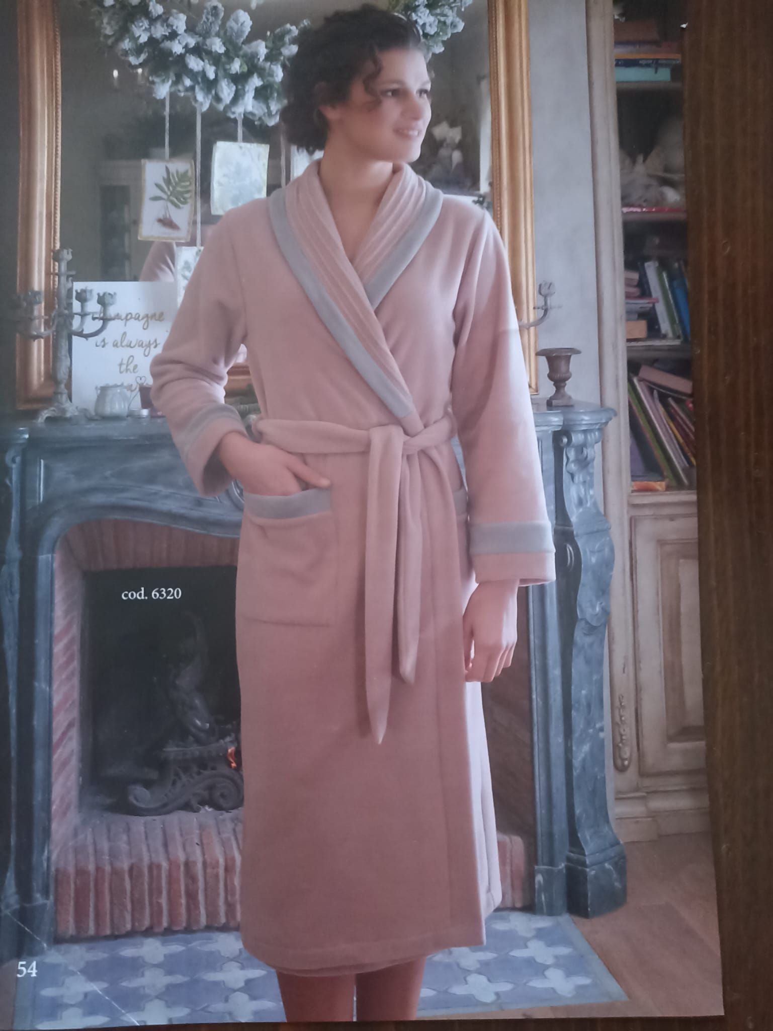 Donna che indossa una lunga tunica rosa con finiture blu, in piedi accanto al camino in una stanza.