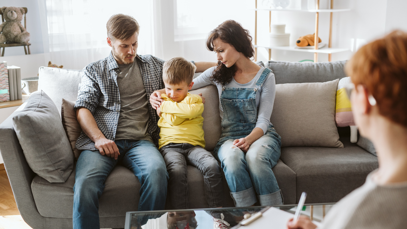 A family in counseling: parents comfort a sad child on a couch; therapist takes notes.