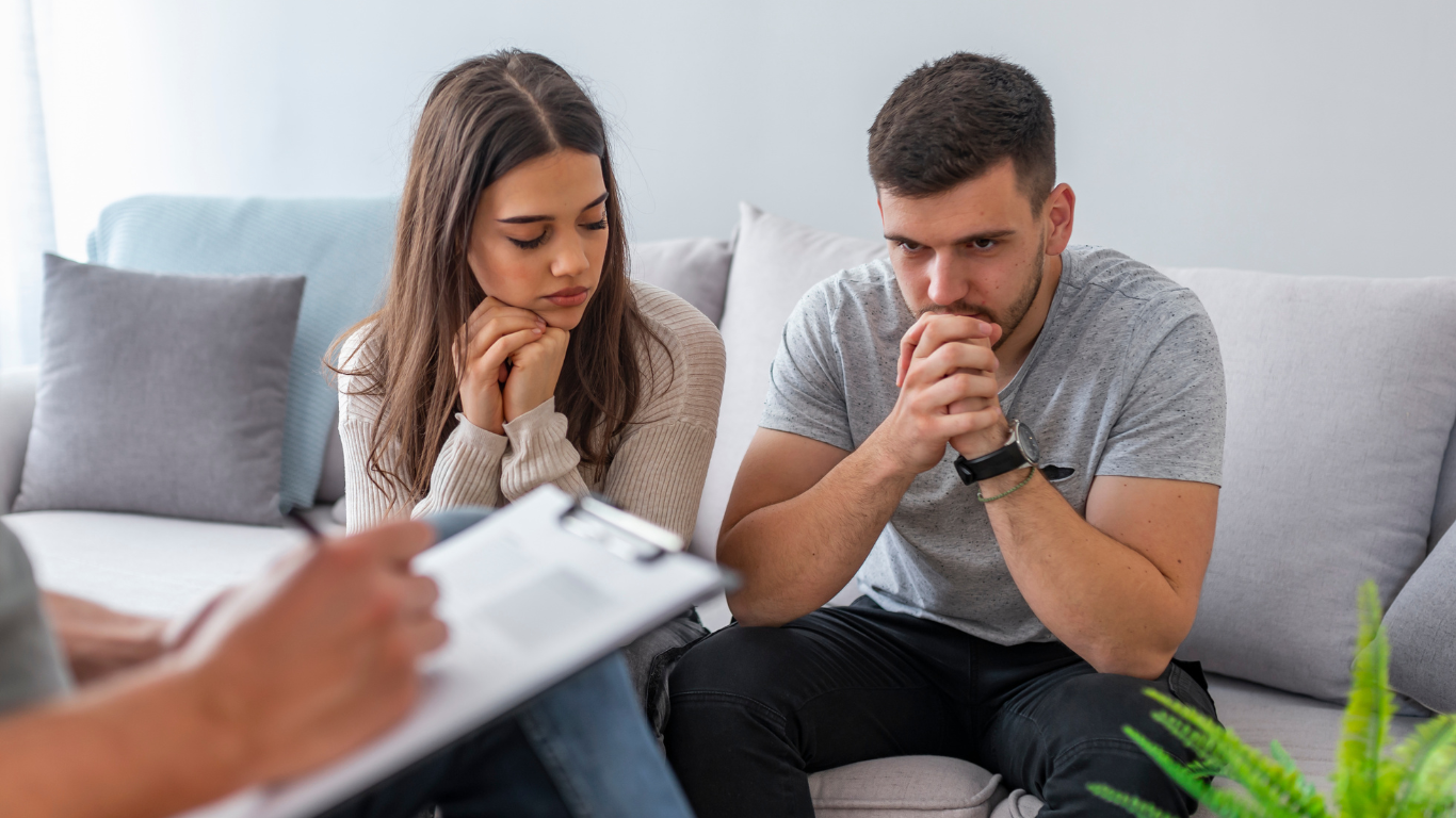 Couple in therapy, looking concerned, seated on a couch while a therapist takes notes.