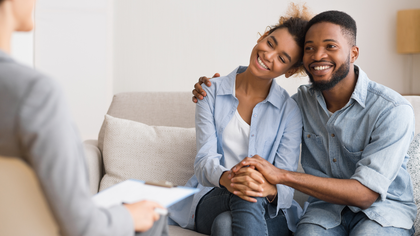 Couple embraces on couch, smiling at therapist in bright room; relationship counseling.