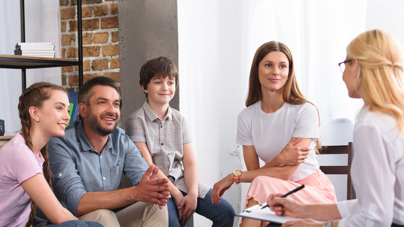 Family in a counseling session, smiling and listening to the therapist, indoors with sunlight.