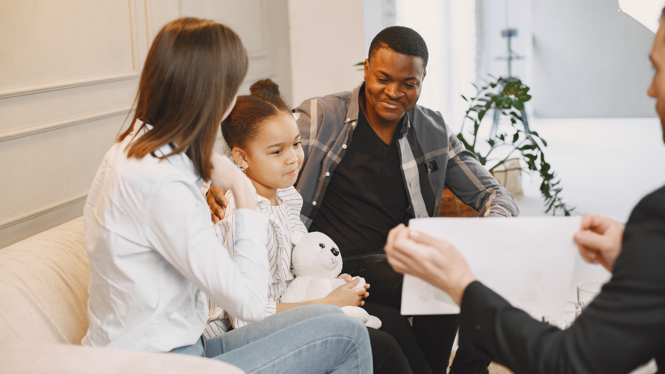 Family, including a young girl, seated on a couch, looking at a document held by a person in a suit; interior, consultation setting. - family counseling