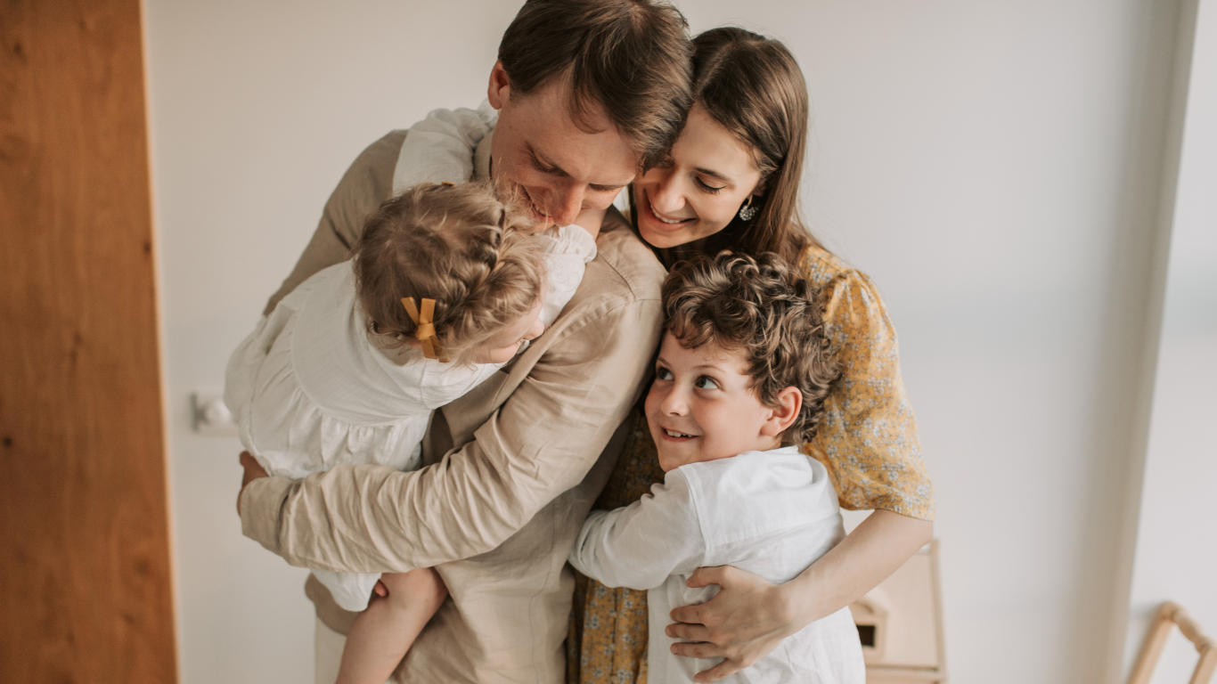 Family of four embracing and smiling; mother and father holding children.