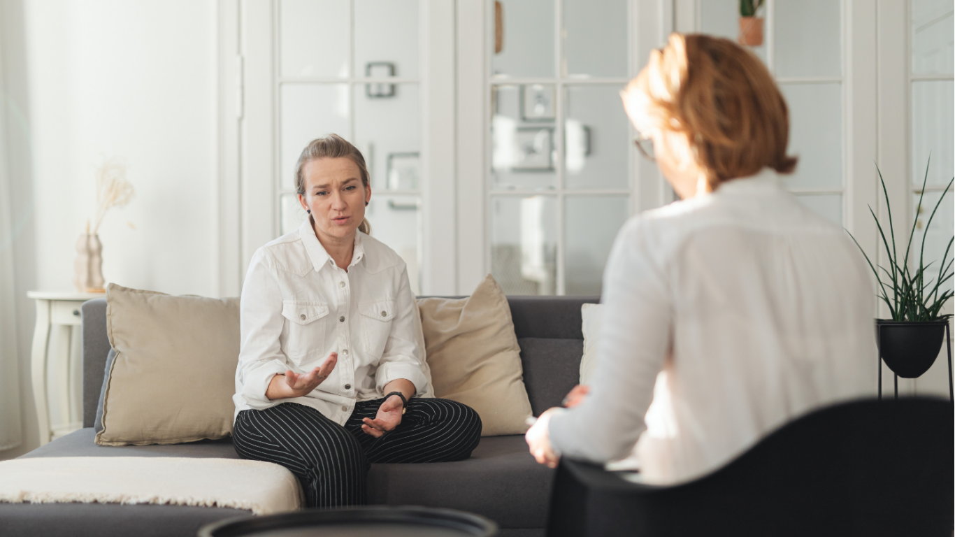 Therapist in glasses takes notes as she listens to a client with red hair.