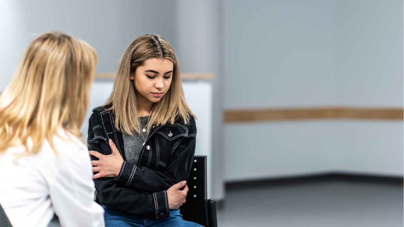 Therapist in glasses takes notes as she listens to a client with red hair.