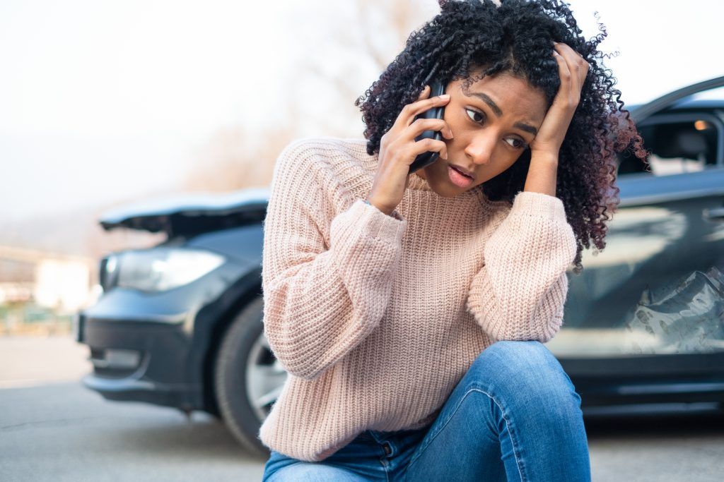 A woman is sitting on the ground talking on a cell phone in front of a damaged car.