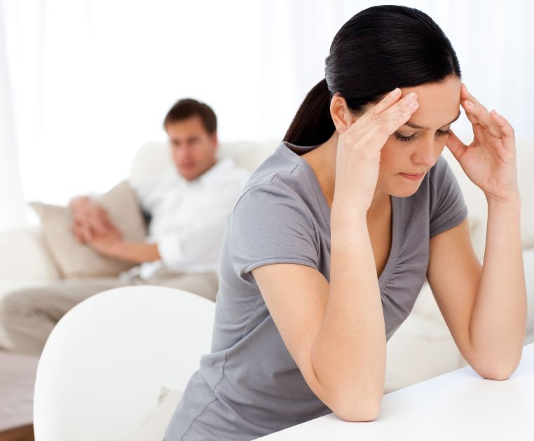 A woman is sitting at a table with her hands on her head while a man sits on a couch in the background