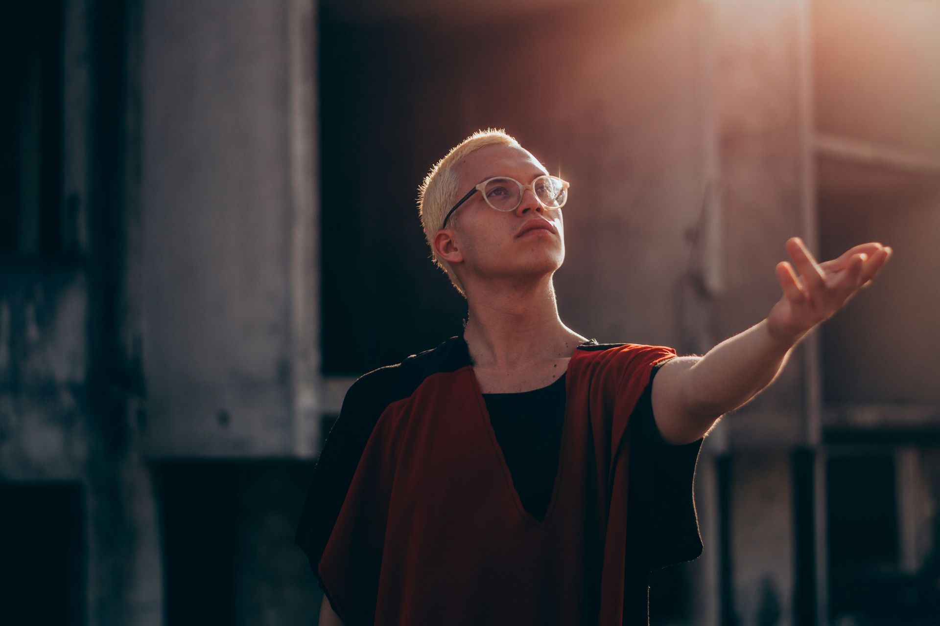 A man wearing glasses is standing in front of a building with his arm outstretched.