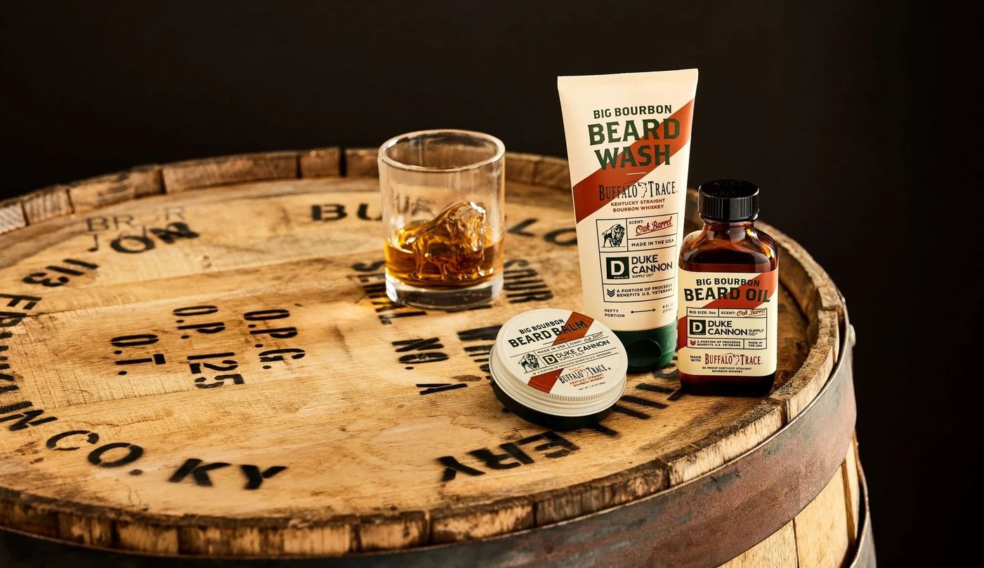 Beard grooming products and a whiskey glass on a wooden barrel.