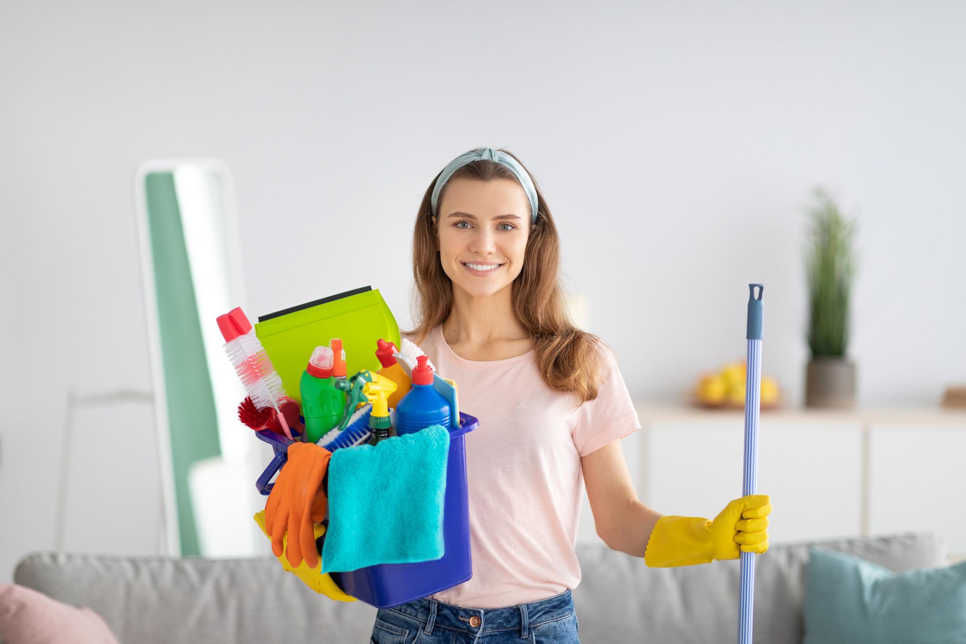 A woman is holding a mop and a basket of cleaning supplies.