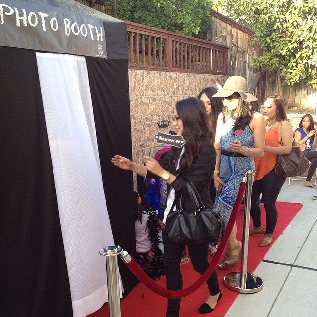 People line up at a photo booth with a red carpet. The woman in the foreground gestures toward the booth.